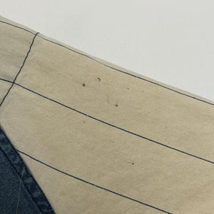 May include: A beige fabric panel featuring thin, evenly spaced vertical black pinstripes. The fabric displays several small, dark brown spots scattered across the surface, indicating minor wear or imperfections on the textile material.