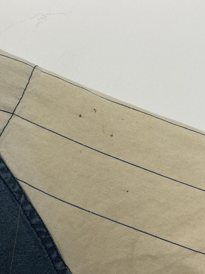 May include: A beige fabric panel featuring thin, evenly spaced vertical black pinstripes. The fabric displays several small, dark brown spots scattered across the surface, indicating minor wear or imperfections on the textile material.