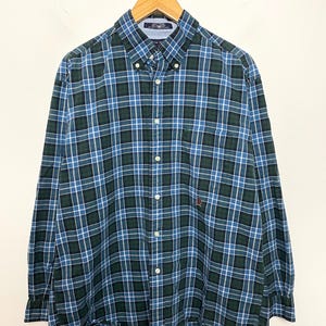Pode incluir: Camisa de manga comprida com padrão xadrez azul, verde e branco. A camisa tem gola abotoada, bolso no peito e um pequeno logótipo bordado. Está pendurada num cabide de madeira.