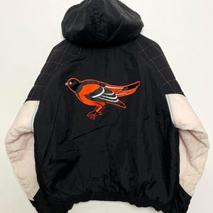 Op de afbeelding: Zwarte capuchontrui met een wit en roze kleurblok ontwerp. De trui heeft een vogellogo op de rug met oranje en zwarte accenten.
