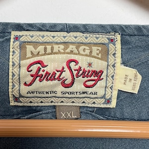 May include: A blue garment featuring a rectangular fabric label with the text Mirage First String Authentic Sportswear in red and black lettering. A smaller tag attached to the side reads Made in Hong Kong, and a brown tag below indicates size XXL.