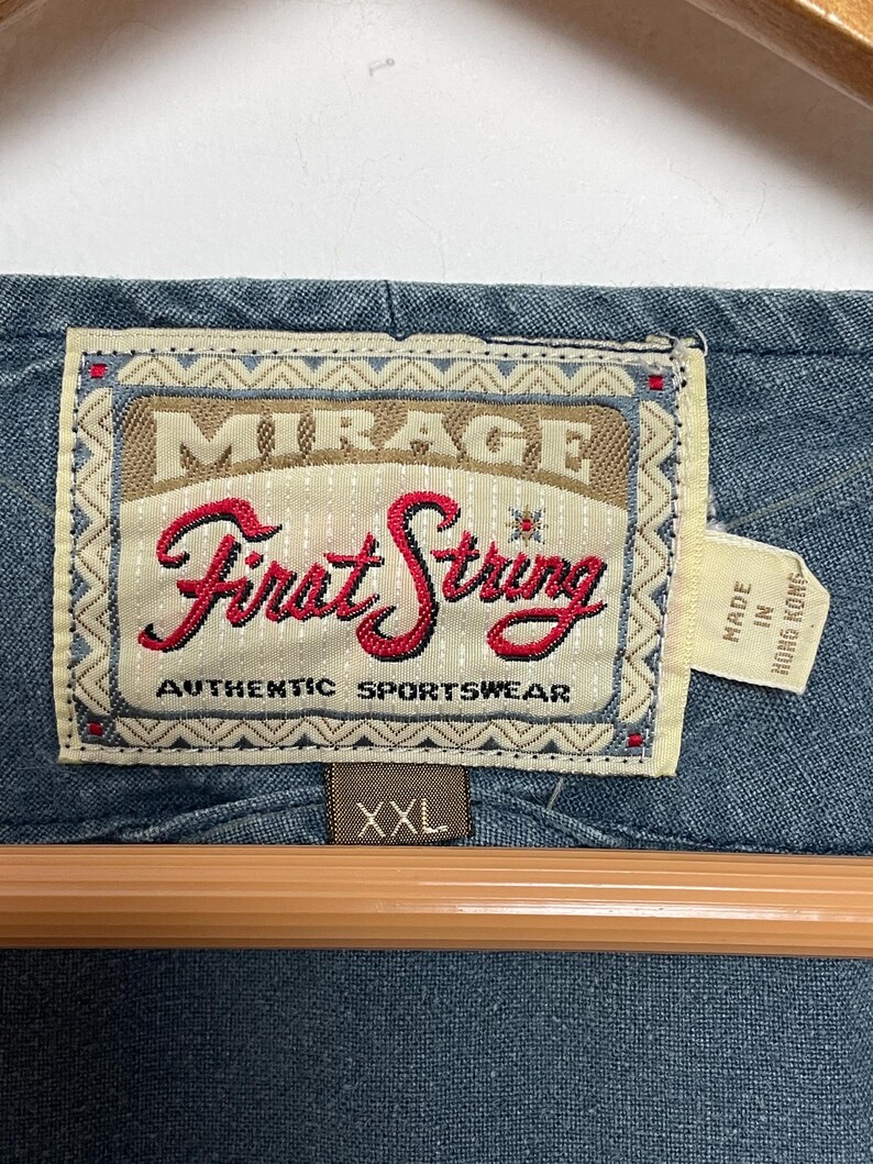 May include: A blue garment featuring a rectangular fabric label with the text Mirage First String Authentic Sportswear in red and black lettering. A smaller tag attached to the side reads Made in Hong Kong, and a brown tag below indicates size XXL.
