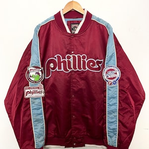 Peut inclure: Une veste de baseball en satin rouge avec des accents bleus et blancs. La veste présente le mot "Phillies" en grandes lettres blanches avec des contours bleus. La veste comporte également plusieurs patchs, dont un patch de la Série mondiale de 1980.