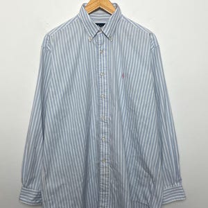 Puede incluir: Camisa abotonada con rayas verticales azules y blancas, colgada de una percha de madera. La camisa tiene mangas largas, cuello abotonado y un pequeño logotipo rosa en el pecho.
