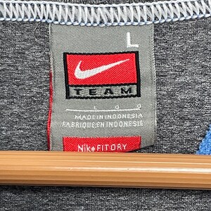 Puede incluir: Primer plano de una camiseta deportiva gris con una etiqueta Nike Team. La etiqueta es rectangular con fondo rojo y un logotipo Nike blanco. El texto "TEAM" es negro. La etiqueta tambi&eacute;n dice "Nike FITDRY".