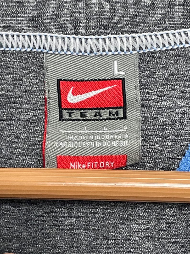 Puede incluir: Primer plano de una camiseta deportiva gris con una etiqueta Nike Team. La etiqueta es rectangular con fondo rojo y un logotipo Nike blanco. El texto "TEAM" es negro. La etiqueta tambi&eacute;n dice "Nike FITDRY".