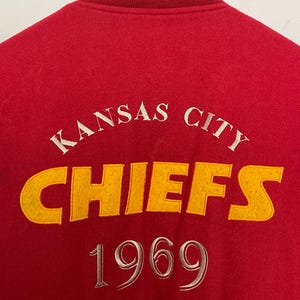 May include: A red varsity jacket with a yellow and gold striped collar. The back displays "KANSAS CITY CHIEFS 1969" in white and yellow. The jacket is made of a soft, textured material. The lettering is embroidered.