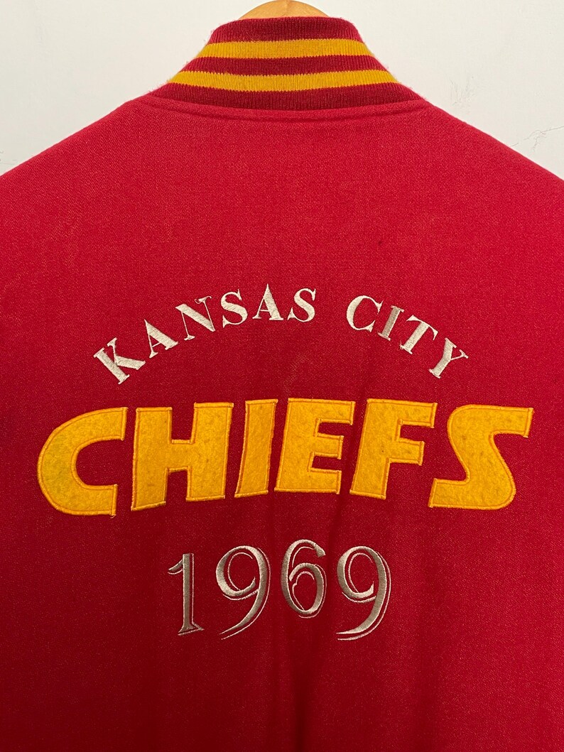 May include: A red varsity jacket with a yellow and gold striped collar. The back displays "KANSAS CITY CHIEFS 1969" in white and yellow. The jacket is made of a soft, textured material. The lettering is embroidered.