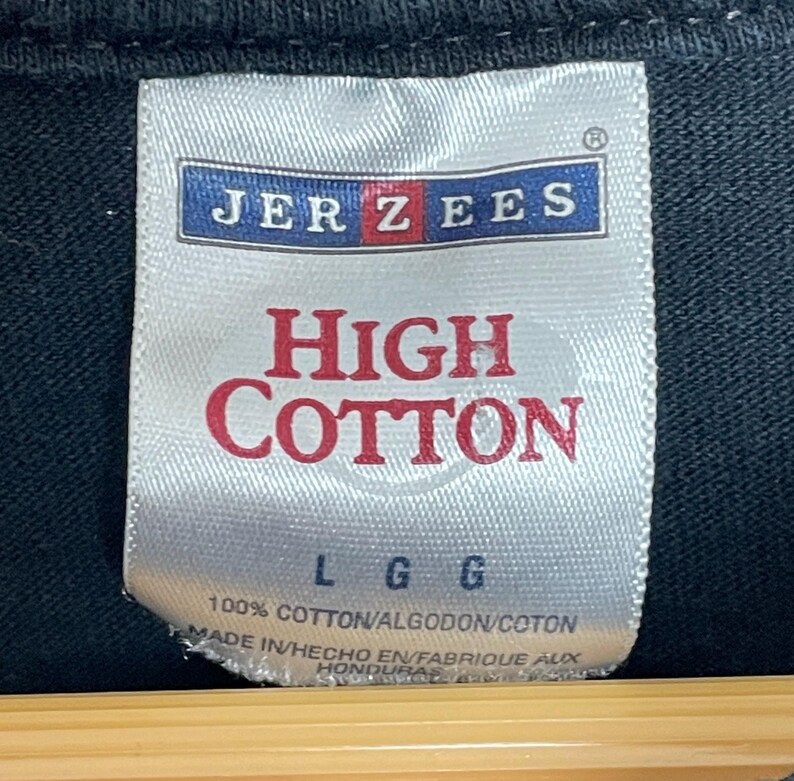May include: Close-up of a white tag with the brand name "JERZEES" in blue and red. The tag reads "HIGH COTTON" in red, with "L G G" below. The bottom of the tag states "100% COTTON" and "MADE IN HONDURAS".