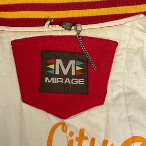 May include: Close-up of a vintage white, red, and gold jacket. The jacket features a red patch with the word "MIRAGE" and a stylized "M" logo. The collar is red and gold striped. The jacket is labeled "XXL" and "Made in Hong Kong."