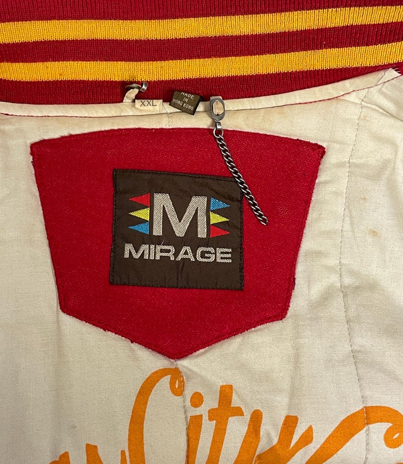 May include: Close-up of a vintage white, red, and gold jacket. The jacket features a red patch with the word "MIRAGE" and a stylized "M" logo. The collar is red and gold striped. The jacket is labeled "XXL" and "Made in Hong Kong."