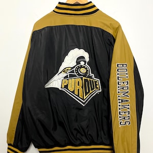 May include: Black and gold Purdue Boilermakers jacket with a train logo and "PURDUE" text. The jacket has gold sleeves and trim, with "BOILERMAKERS" printed on the right sleeve. The jacket is hanging on a wooden hanger.