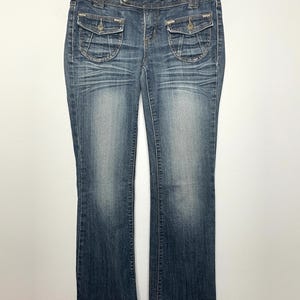 May include: Blue denim jeans with a faded wash. The jeans have two front pockets with button closures and two back pockets. The jeans are hanging on a wooden hanger.