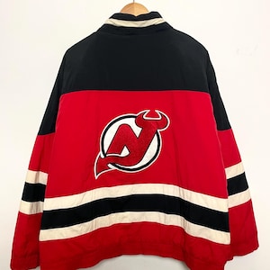 May include: A red, black, and white jacket with the New Jersey Devils logo on the back. The jacket has a black upper section, a red midsection with the logo, and red sleeves with black and white stripes.