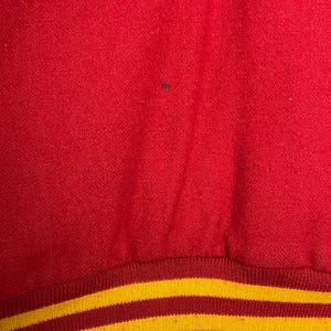 May include: Close-up of red fabric with a textured surface. The bottom edge has a ribbed band with two yellow stripes on a red background. The fabric has a small dark spot.