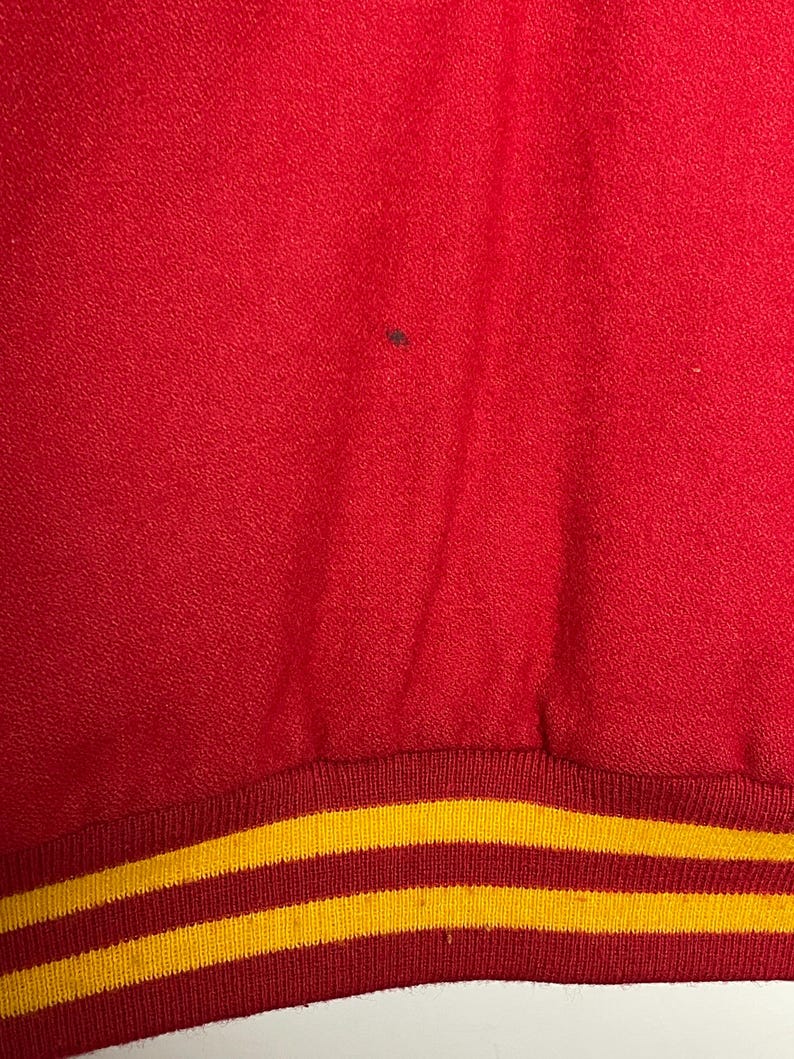 May include: Close-up of red fabric with a textured surface. The bottom edge has a ribbed band with two yellow stripes on a red background. The fabric has a small dark spot.