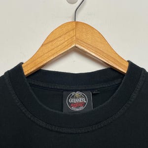 May include: Black crew neck t-shirt on a wooden hanger. The shirt displays a circular Guinness label with the words "Official Merchandise". The hanger is suspended from a white wall. The t-shirt is a classic style.