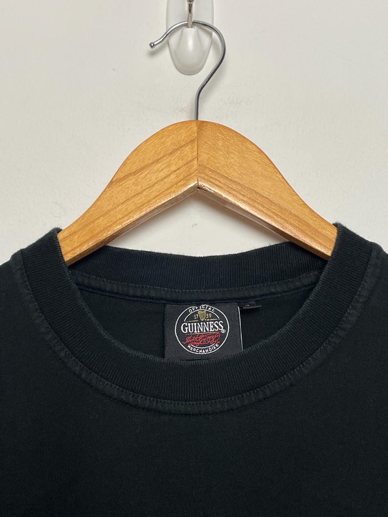 May include: Black crew neck t-shirt on a wooden hanger. The shirt displays a circular Guinness label with the words "Official Merchandise". The hanger is suspended from a white wall. The t-shirt is a classic style.
