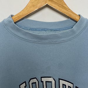 K&ouml;nnte beinhalten: Ein hellblauer Pullover mit einem wei&szlig;en und schwarzen Grafikdruck des Wortes "FORD" in gro&szlig;er, fetter Schrift.
