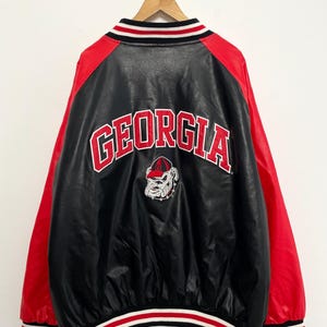 Puede incluir: Chaqueta universitaria negra y roja con la palabra "GEORGIA" en letras rojas en la espalda. La chaqueta presenta un logotipo de bulldog y un cuello y puños a rayas. Las mangas son rojas y el cuerpo es negro.