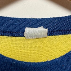 K&ouml;nnte beinhalten: Nahaufnahme eines blau-gelb gestreiften T-Shirts. Das Shirt hat einen blauen Kragen und ein wei&szlig;es Etikett am Ausschnitt. Der Stoff scheint Baumwolle oder ein &auml;hnliches Material zu sein.