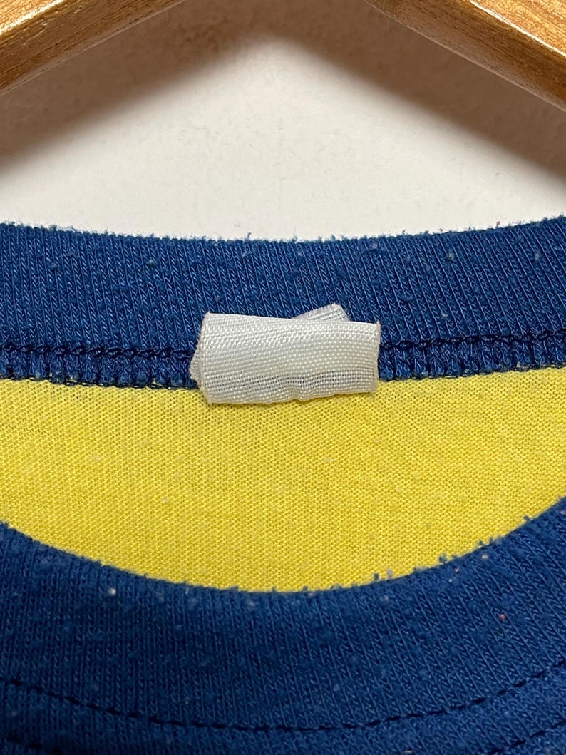 K&ouml;nnte beinhalten: Nahaufnahme eines blau-gelb gestreiften T-Shirts. Das Shirt hat einen blauen Kragen und ein wei&szlig;es Etikett am Ausschnitt. Der Stoff scheint Baumwolle oder ein &auml;hnliches Material zu sein.