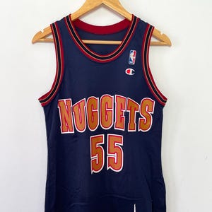 Può includere: Una maglia da basket blu navy con la scritta "Nuggets" e il numero "55" in lettere dorate con contorni rossi. La maglia ha un bordo rosso, nero e oro intorno al collo e alle ascelle. Il logo NBA e un logo Champion sono sul davanti della maglia.