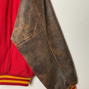 May include: A vintage varsity jacket in red and brown. The jacket has a red body and brown leather sleeves. The cuffs and waistband feature yellow and red stripes. The leather sleeves show signs of wear, giving it a vintage look.