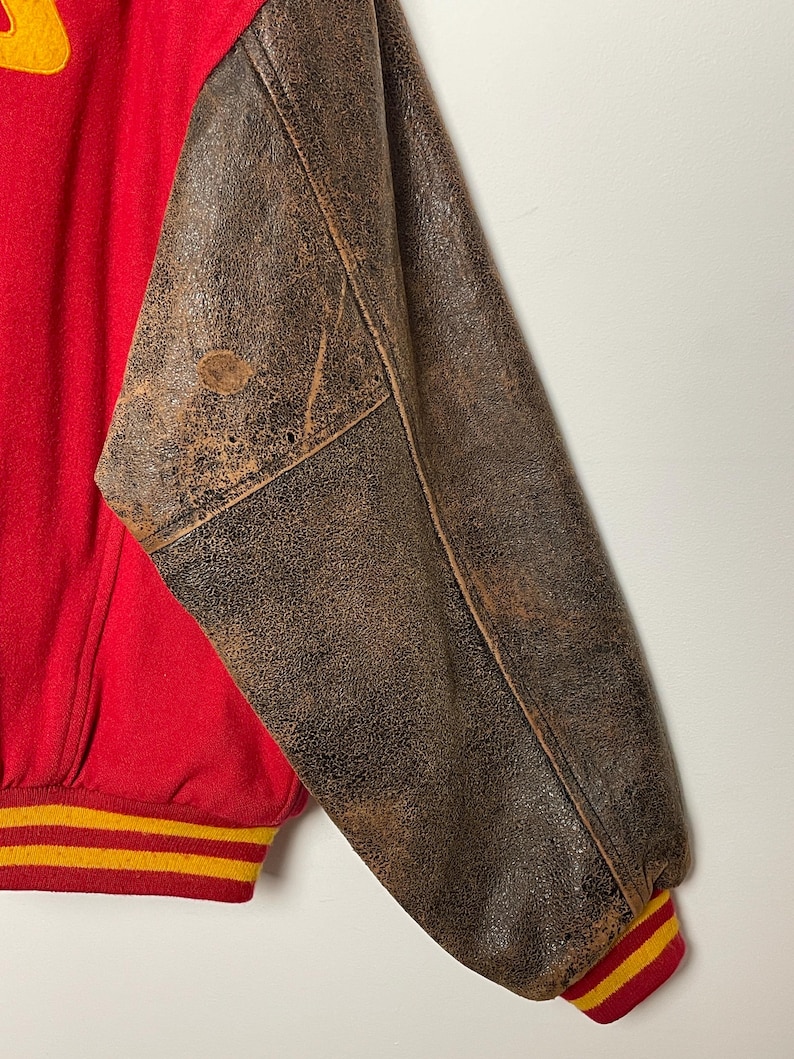 May include: A vintage varsity jacket in red and brown. The jacket has a red body and brown leather sleeves. The cuffs and waistband feature yellow and red stripes. The leather sleeves show signs of wear, giving it a vintage look.