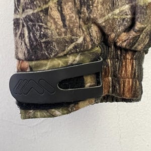 Pode incluir: Close-up de um tecido com padr&atilde;o de camuflagem com um clip preto. O tecido apresenta tons de castanho, verde e bege. O clip &eacute; fixado ao tecido com um fecho de velcro.