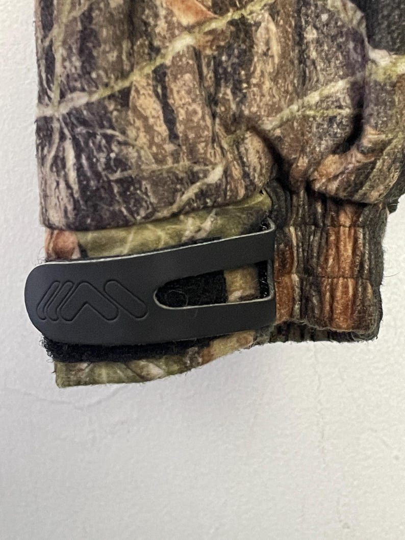 Pode incluir: Close-up de um tecido com padr&atilde;o de camuflagem com um clip preto. O tecido apresenta tons de castanho, verde e bege. O clip &eacute; fixado ao tecido com um fecho de velcro.
