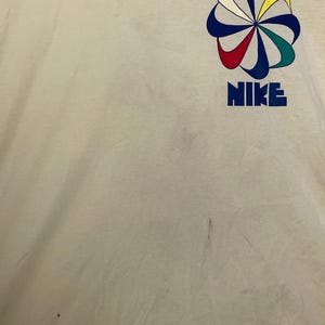 May include: Cream-coloured t-shirt with a colourful graphic design and the word "NIKE" in blue block letters. The graphic is a stylised flower with red, yellow, green, and blue petals.