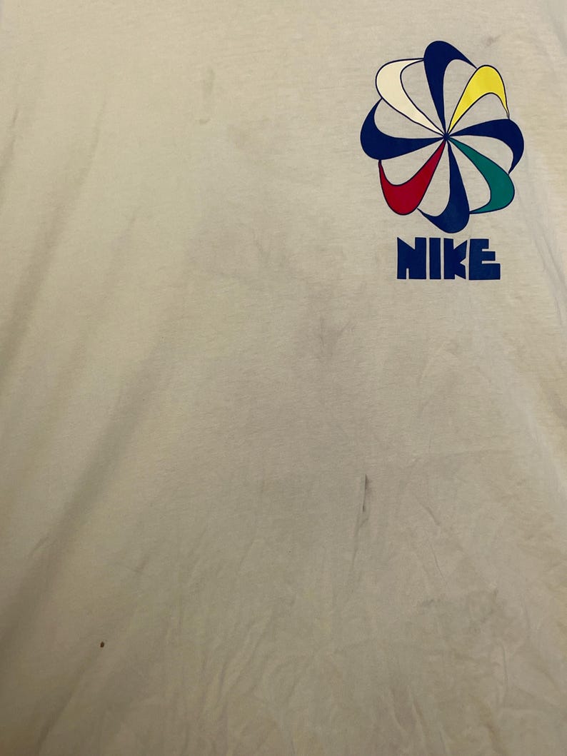 May include: Cream-coloured t-shirt with a colourful graphic design and the word "NIKE" in blue block letters. The graphic is a stylised flower with red, yellow, green, and blue petals.