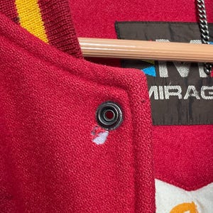 May include: Close-up of a red varsity jacket with a yellow and gold striped collar. The jacket has a snap closure and a label that reads "MIRAGE". The jacket is made of a textured material.