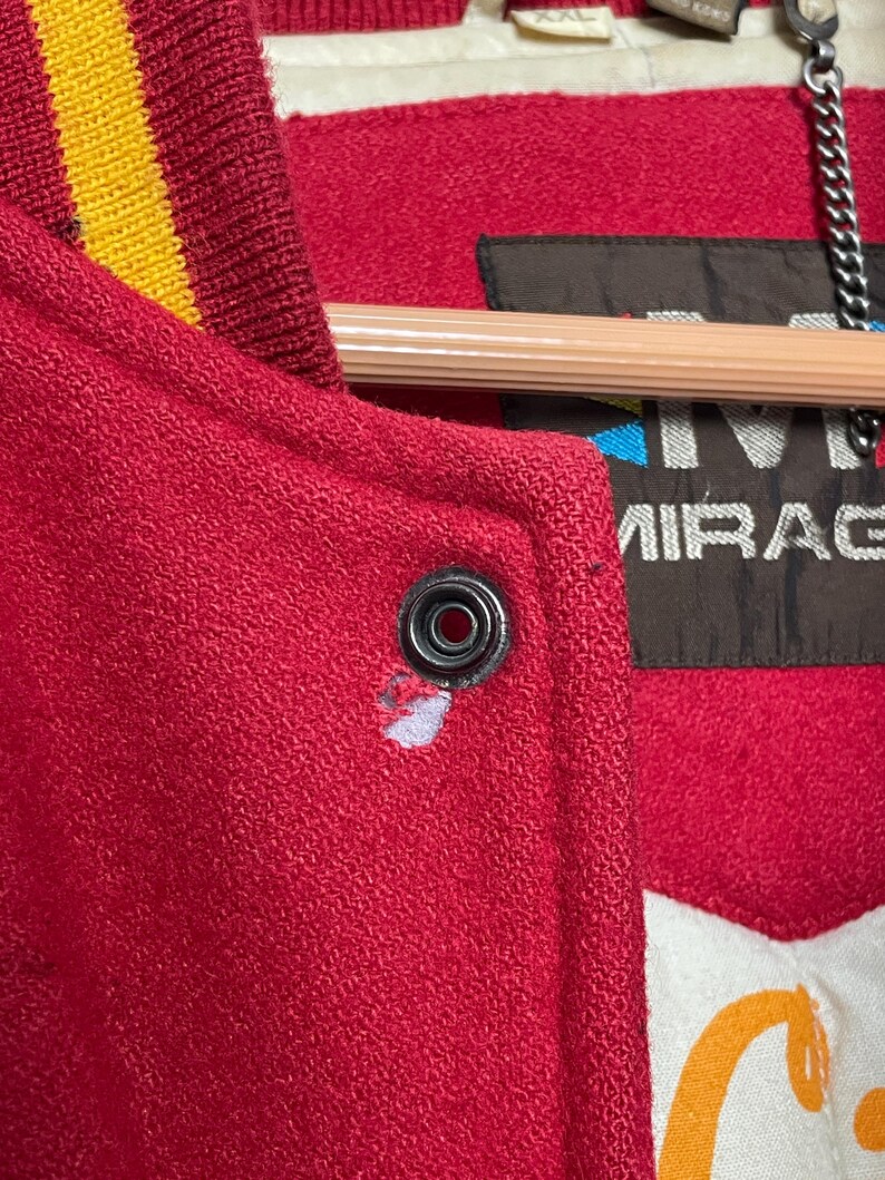 May include: Close-up of a red varsity jacket with a yellow and gold striped collar. The jacket has a snap closure and a label that reads "MIRAGE". The jacket is made of a textured material.