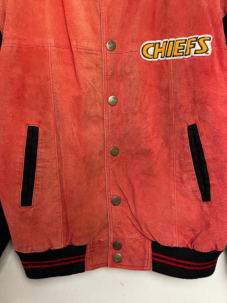 Puede incluir: Chaqueta bomber de cuero roja con pu&ntilde;os a rayas rojas y negras y un parche "CHIEFS" en blanco y negro en el pecho.