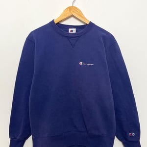 May include: A navy blue Champion sweatshirt with a crew neck and long sleeves. The Champion logo is embroidered in white and red on the chest and left sleeve cuff. The sweatshirt is hanging on a wooden hanger.