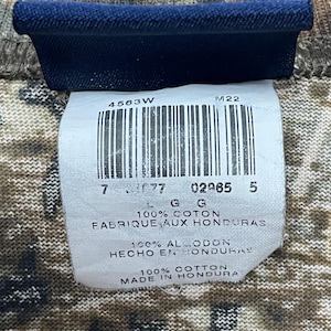 May include: A close-up shot of a clothing label with a barcode and text. The label is white with black text and a blue border. The text includes "100% COTTON" and "MADE IN HONDURAS". The background is a camouflage pattern.