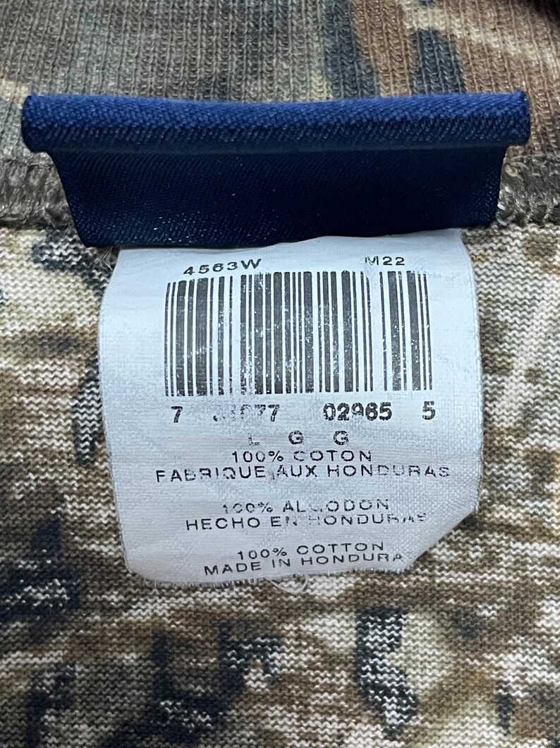May include: A close-up shot of a clothing label with a barcode and text. The label is white with black text and a blue border. The text includes "100% COTTON" and "MADE IN HONDURAS". The background is a camouflage pattern.