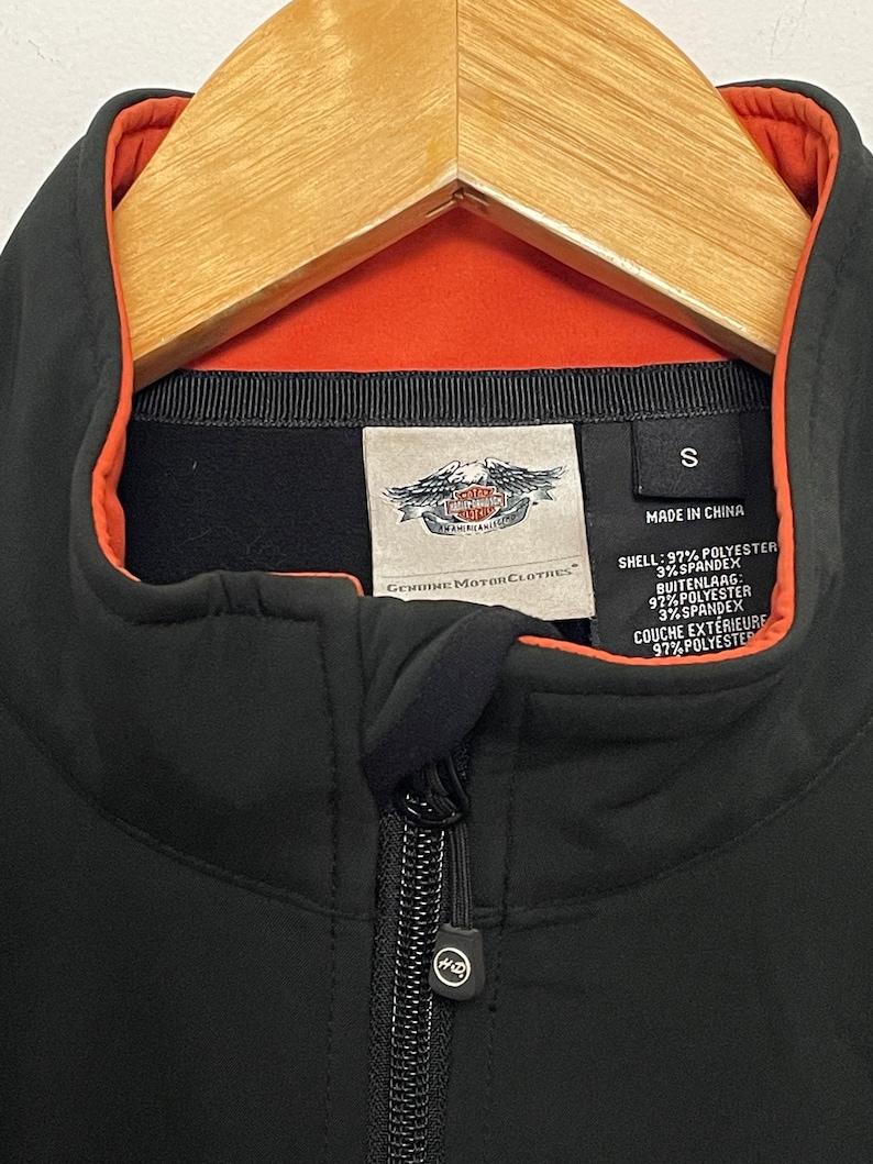 May include: Black zippered jacket with an orange lining and a Harley Davidson logo. The tag says "Genuine Motor Clothes" and lists the fabric composition as 97% polyester and 3% spandex.