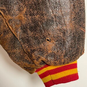 May include: Close-up of a vintage brown leather jacket with a distressed, textured appearance. The jacket's sleeve cuff features red, yellow, and white horizontal stripes. A small hole is visible in the leather.