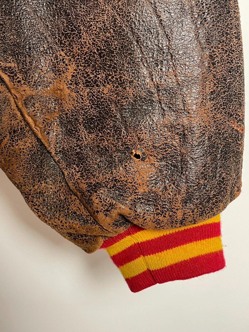 May include: Close-up of a vintage brown leather jacket with a distressed, textured appearance. The jacket's sleeve cuff features red, yellow, and white horizontal stripes. A small hole is visible in the leather.