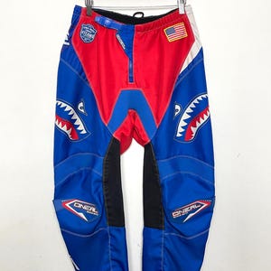 May include: Blue and red motocross pants with a shark graphic and the O'Neal logo. The pants have a black waistband and a zipper fly. The pants also have a small American flag patch on the right leg.