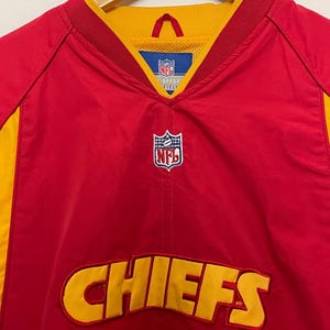 Puede incluir: Chaqueta de entrenamiento roja y amarilla de los Chiefs de la NFL. La chaqueta presenta el nombre del equipo "CHIEFS" en letras amarillas en la parte delantera. El logotipo de la NFL es visible en el pecho y el cuello. La chaqueta est&aacute; colgada en una percha de madera.