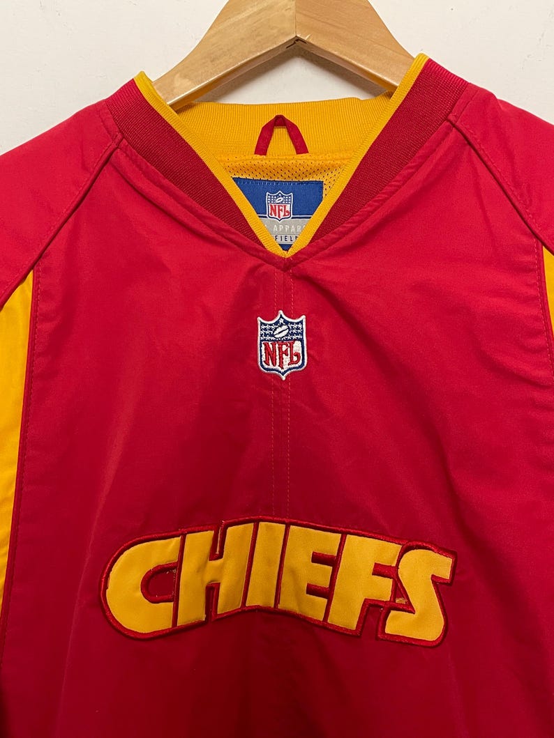 Puede incluir: Chaqueta de entrenamiento roja y amarilla de los Chiefs de la NFL. La chaqueta presenta el nombre del equipo "CHIEFS" en letras amarillas en la parte delantera. El logotipo de la NFL es visible en el pecho y el cuello. La chaqueta est&aacute; colgada en una percha de madera.