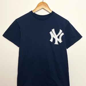 Può includere: T-shirt blu navy con logo bianco dei New York Yankees sul petto. Il logo è un design "NY" ad incastro. La maglietta ha maniche corte e girocollo. Un capo d'abbigliamento sportivo classico.