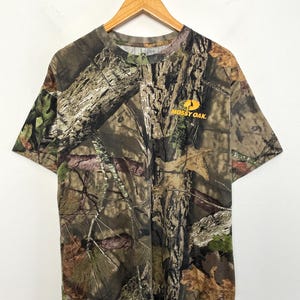 May include: A short-sleeved t-shirt with a camouflage pattern in shades of brown, green, and tan. The shirt features the "Mossy Oak" logo in orange on the right chest. The shirt is hanging on a wooden hanger.
