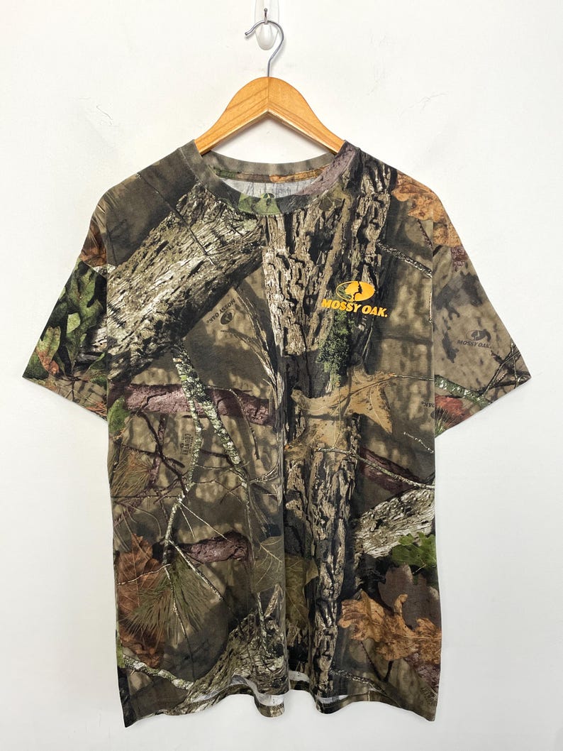 May include: A short-sleeved t-shirt with a camouflage pattern in shades of brown, green, and tan. The shirt features the "Mossy Oak" logo in orange on the right chest. The shirt is hanging on a wooden hanger.
