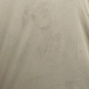 May include: Close-up of a light beige t-shirt with several dark spots. The fabric appears to be slightly wrinkled, with visible folds and creases. The overall texture of the shirt is soft and likely made of cotton or a similar material.