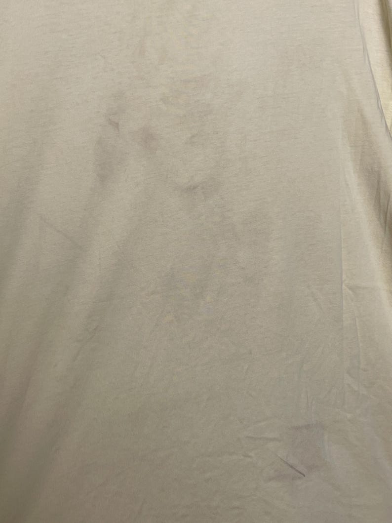 May include: Close-up of a light beige t-shirt with several dark spots. The fabric appears to be slightly wrinkled, with visible folds and creases. The overall texture of the shirt is soft and likely made of cotton or a similar material.